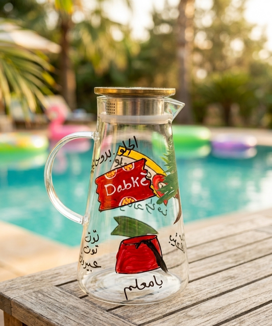 Hand-painted glass pitcher with Lebanese heritage motifs sitting on a wooden table by a pool